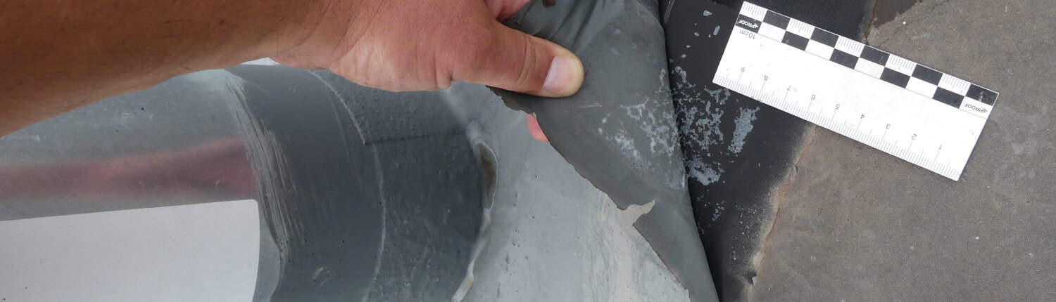 Schadensprüfung an einem Dachanschluss Detailaufnahme einer manuellen Prüfung einer Dachbahn, bei der die Ablösung der Abdichtung mithilfe eines Maßstabs dokumentiert wird.