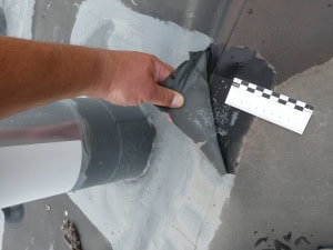 Schadensprüfung an einem Dachanschluss Detailaufnahme einer manuellen Prüfung einer Dachbahn, bei der die Ablösung der Abdichtung mithilfe eines Maßstabs dokumentiert wird.